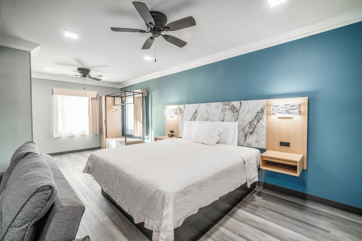 Habitación de hotel moderna con una cama grande, ventilador de techo, mesita de noche con lámpara y vistas al exterior.
