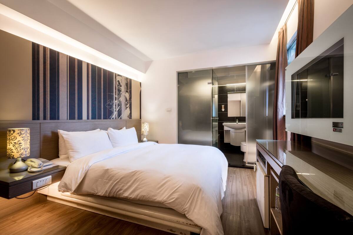A modern hotel room with a large bed, bedside table with a lamp, and a TV.
