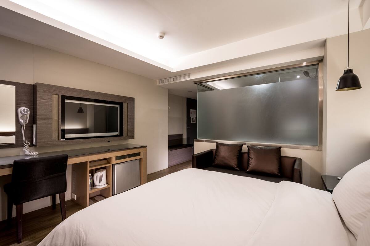 A modern hotel room with a large bed, a desk with a chair, a television, and a large window.