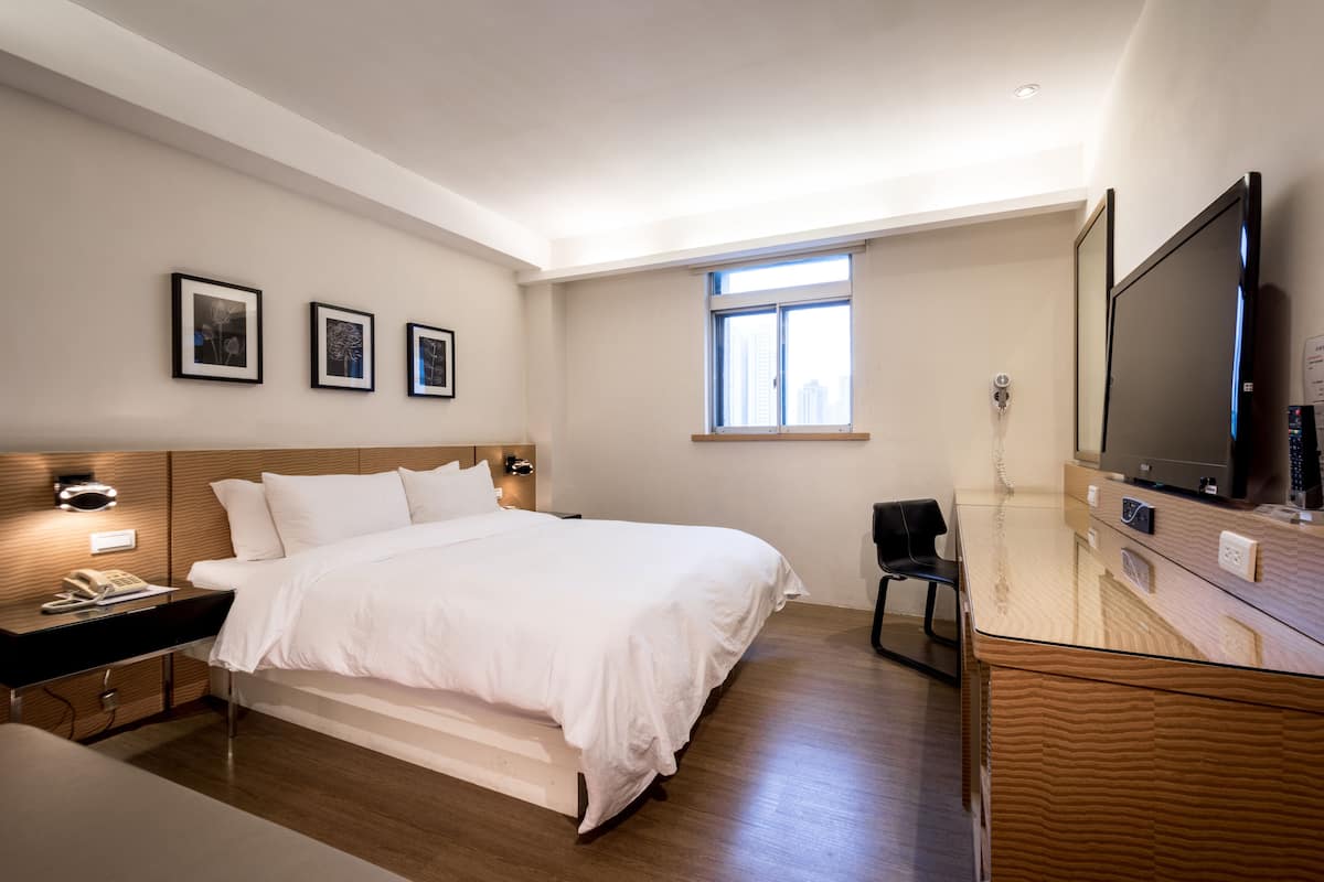 A modern hotel room with a large bed, bedside tables, a desk with a flat-screen TV, and a window with a city view.