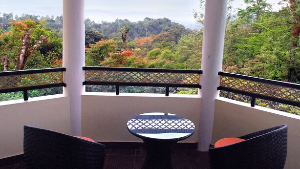 A balcony with a round table and two chairs, overlooking a lush forested area. - Amritasthanam Guest House And Retreat (Madikeri)