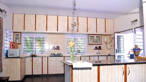 A kitchen with wooden cabinets, a central island, a microwave, and a vase with flowers. - Amritasthanam Guest House And Retreat (Madikeri)