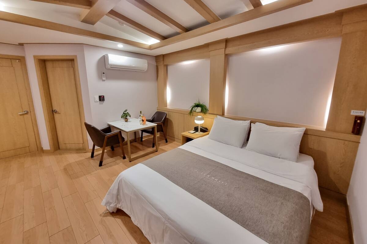 A hotel room with a bed, bedside table, lamp, desk with chair, and an air conditioning unit.