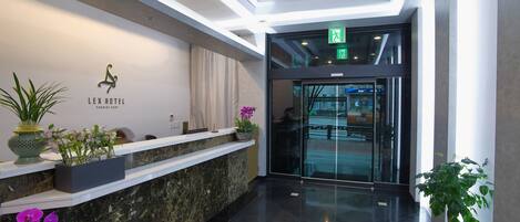 Zona de recepción de hotel con mostrador de mármol, plantas en macetas y puerta de cristal que da al vestíbulo.