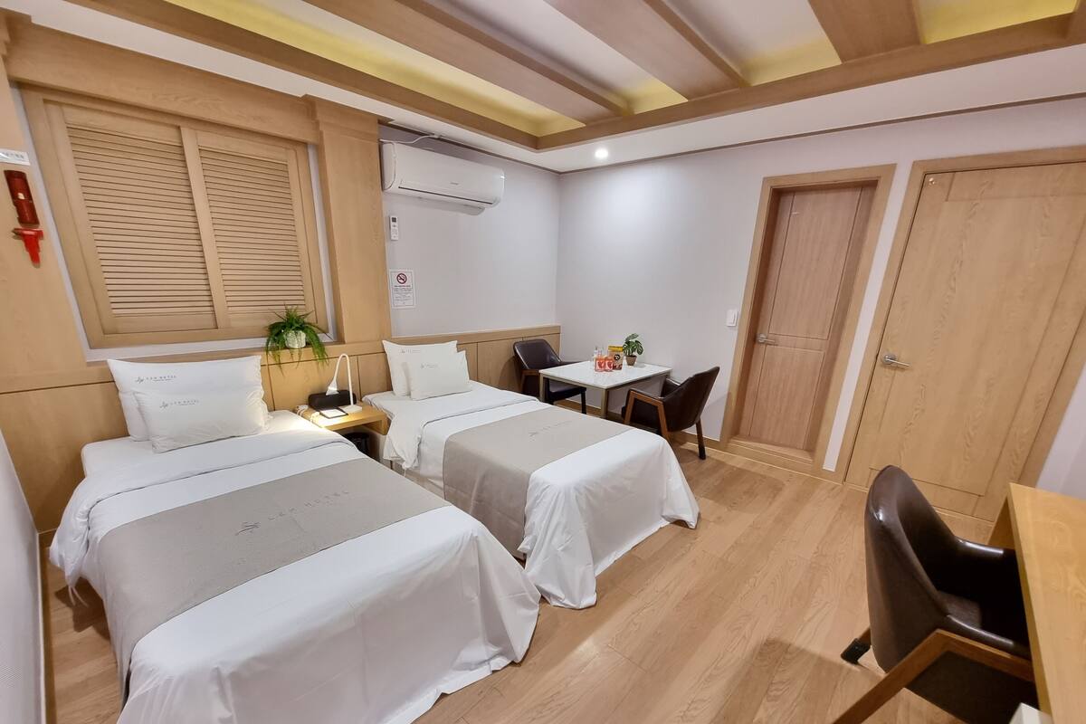 A hotel room with two beds, a desk, a chair, a table, an air conditioning unit, and a door.