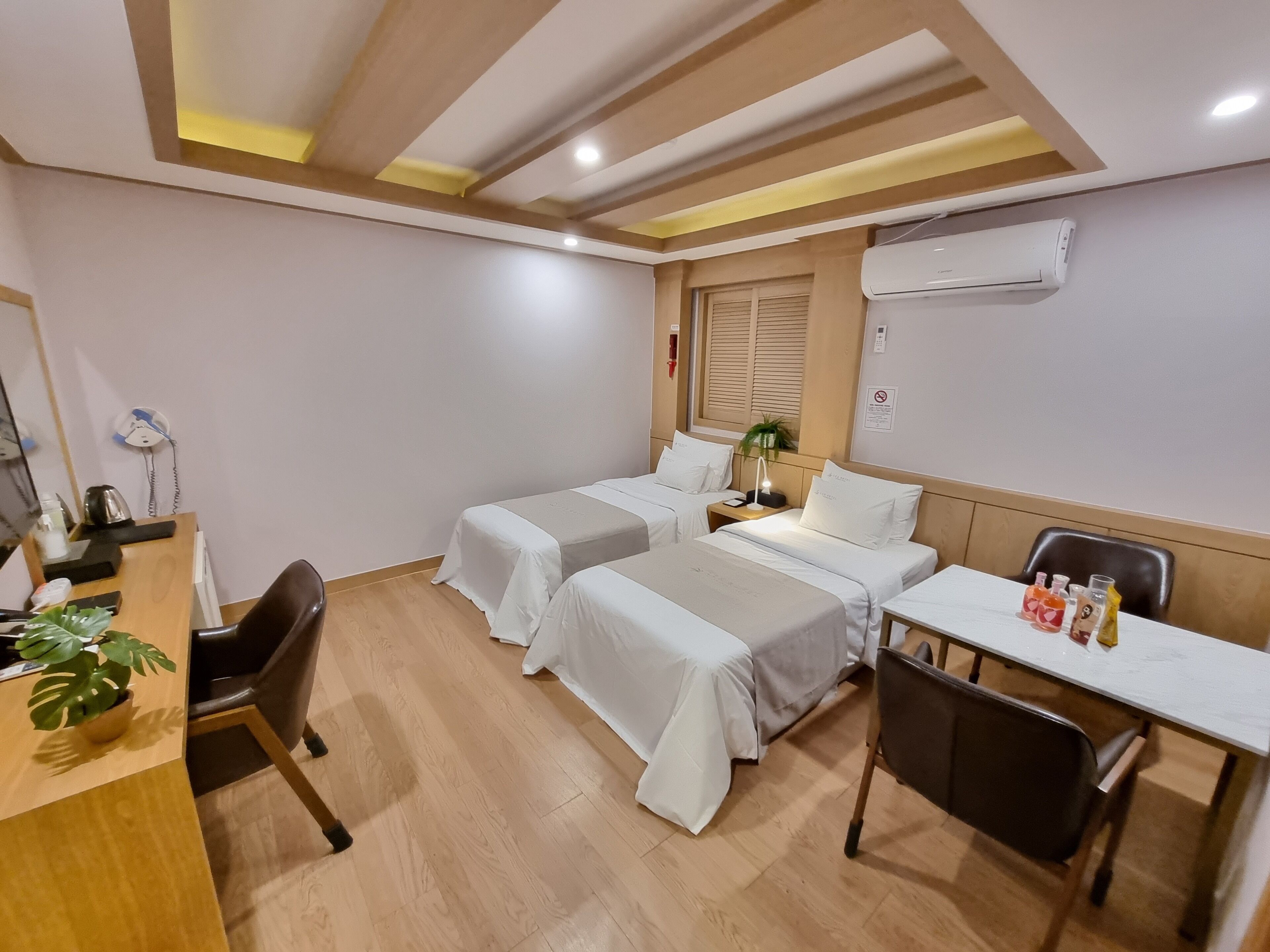 A hotel room with two beds, a desk, a chair, a plant, and an air conditioning unit.
