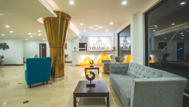 A modern lobby with a reception desk, a blue armchair, a grey sofa, and a decorative pillar. - Vinayaga by Poppys Rameshwaram (Rameshwaram)