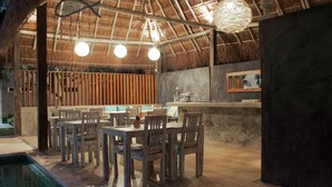 An outdoor dining area with wooden tables and chairs, a pool, and a thatched roof structure. - Villas H2O (Tulum)