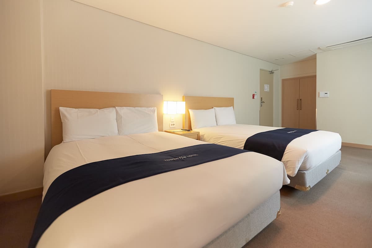 Habitación de hotel con dos camas, cada una con ropa de cama blanca y detalles en azul marino. Un mesita de noche con lámpara se encuentra entre las camas.