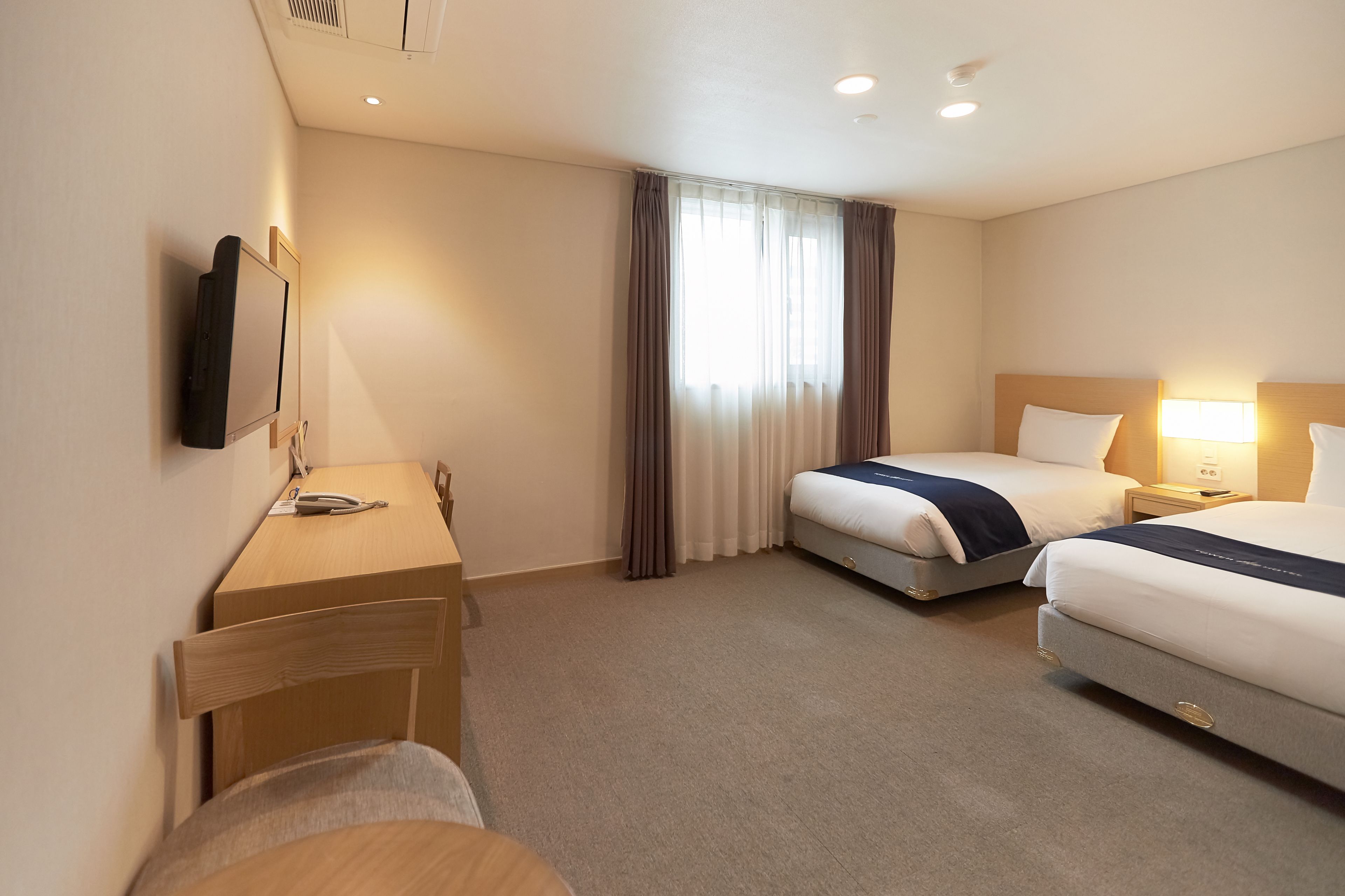 A hotel room with two beds, a desk, a chair, a TV mounted on the wall, and a window with curtains.