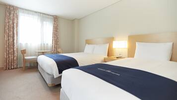 A hotel room with two beds, a small table, a chair, and a window with curtains.