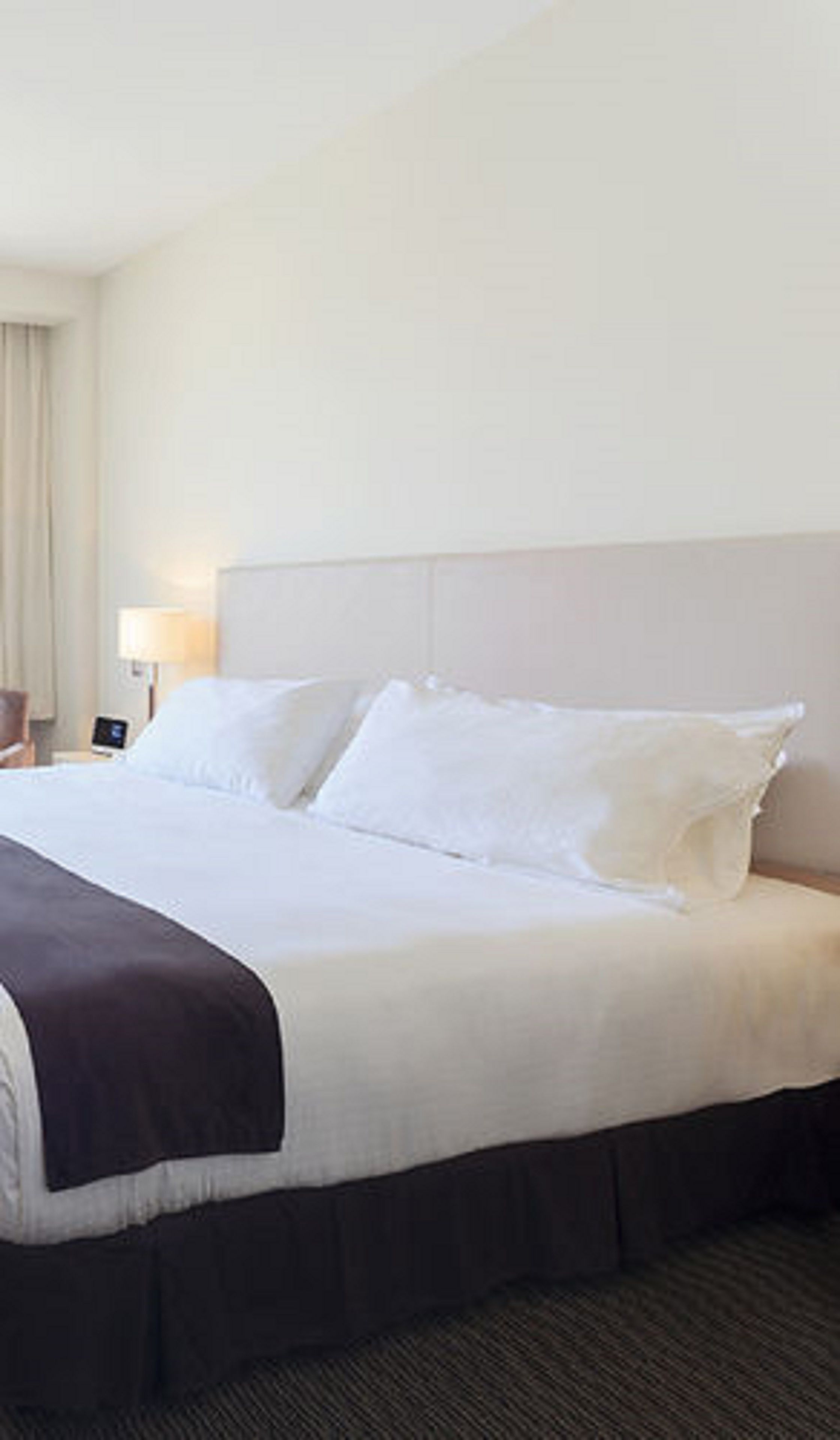 Una habitación de hotel con una cama grande, una mesita de noche, una lámpara y un ventanal con cortinas.