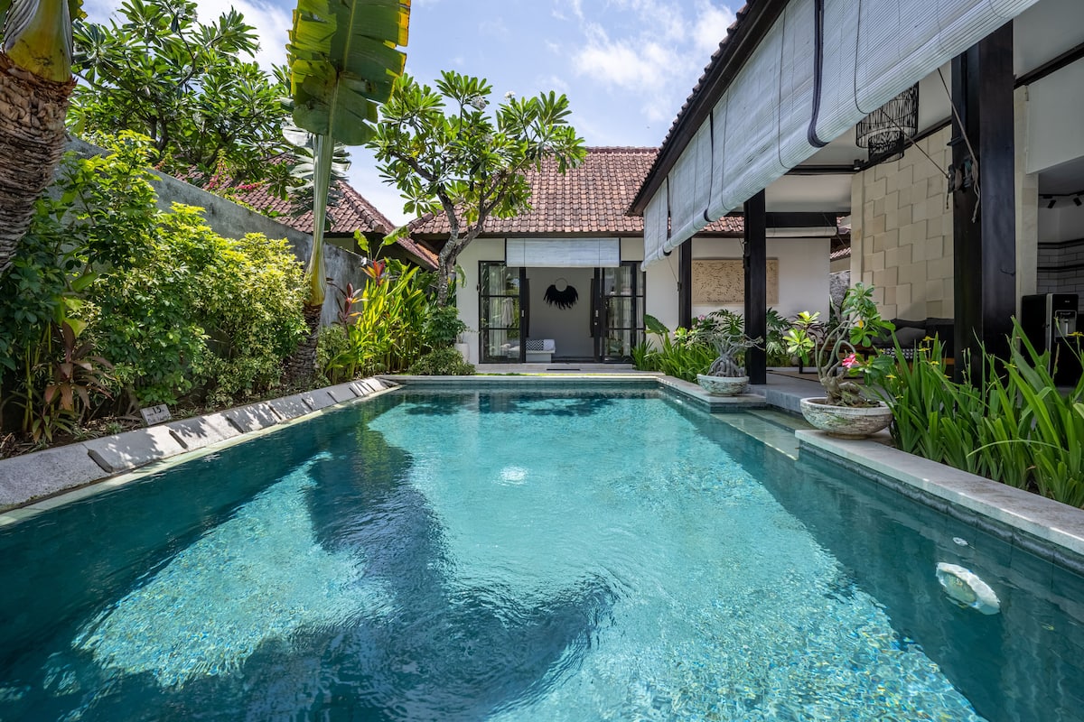 A swimming pool with a tiled deck, surrounded by lush greenery and a building with a tiled roof.
