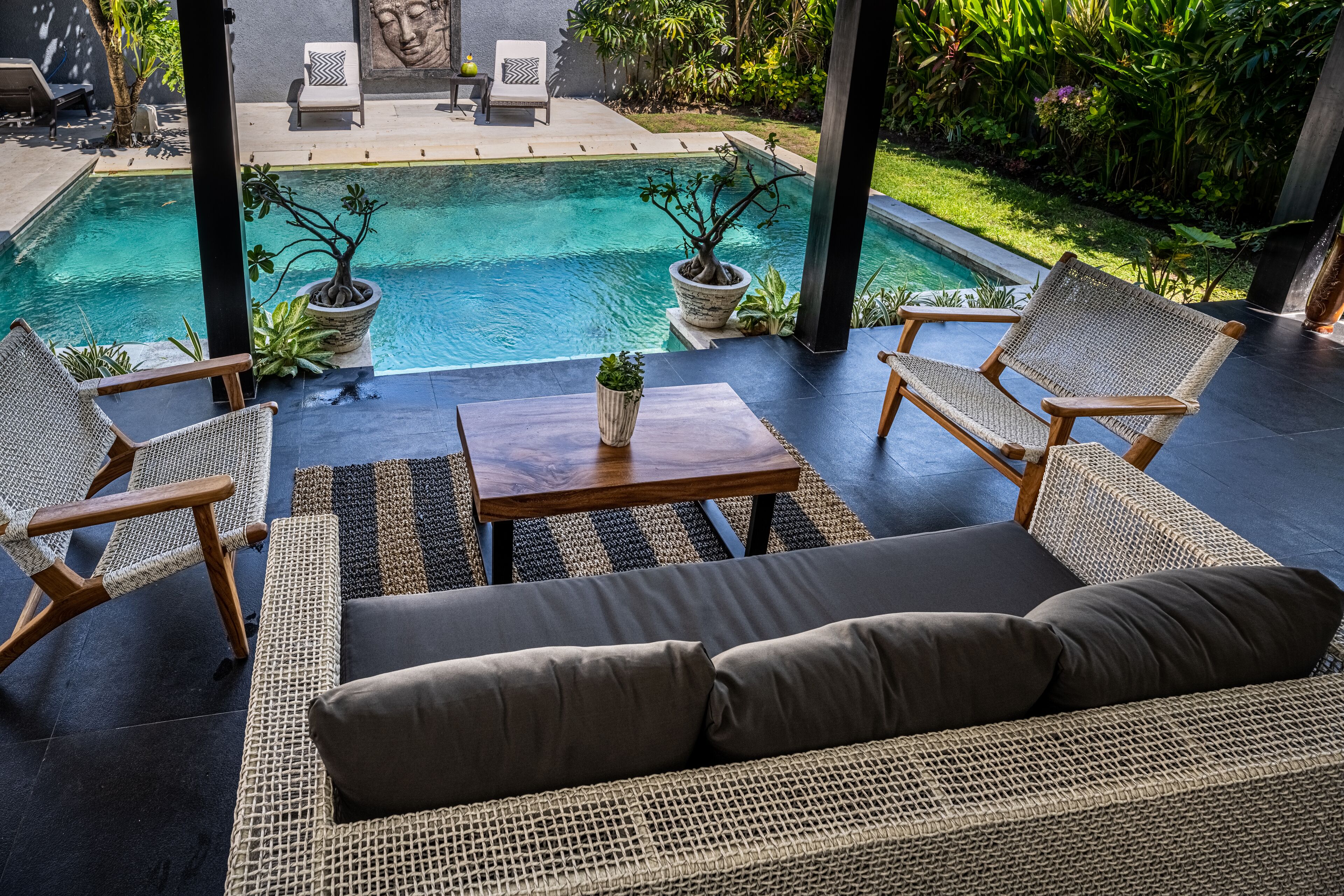 Un área junto a la piscina con mobiliario de mimbre, una mesa de madera y plantas en macetas.