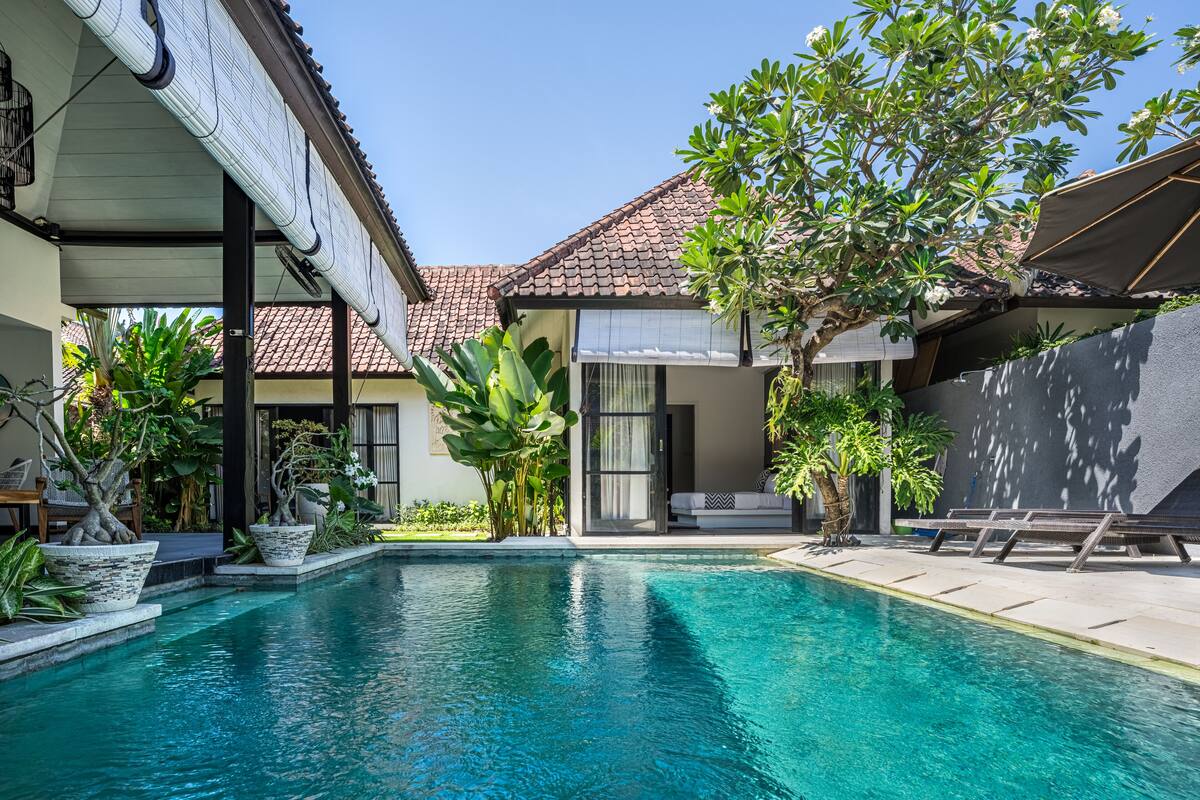 A swimming pool with a tiled deck, surrounded by potted plants and a covered seating area.