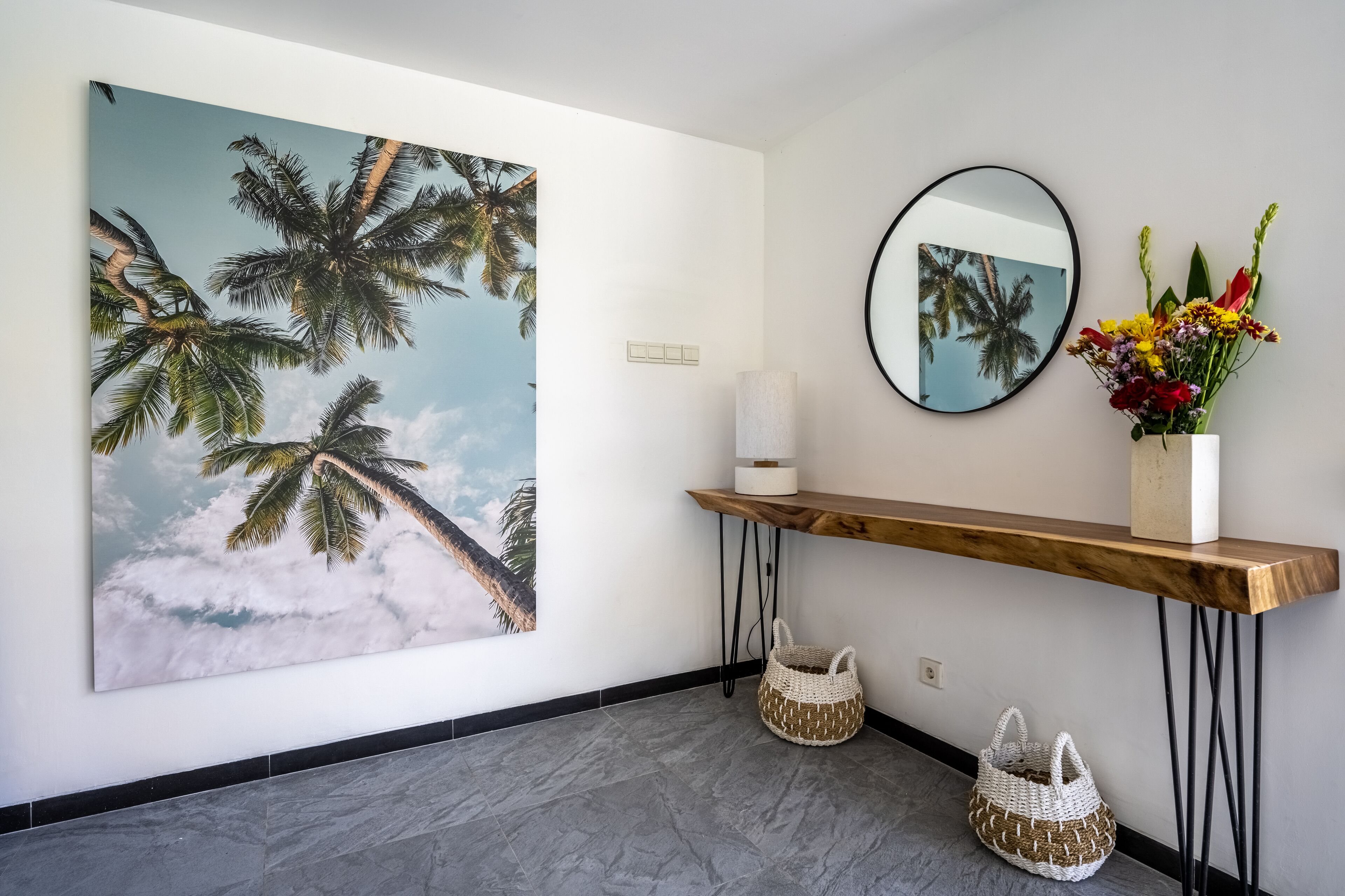 A hallway with a large tropical-themed painting, a mirror, a wooden console table with a vase of flowers, and two woven baskets.