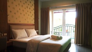 A hotel room with a bed, a nightstand, a window with curtains, and a view of the outside balcony. - Phuket Ecozy Hotel (Wichit)