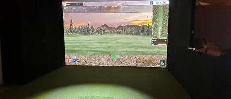 Configuración de simulación de golf con pantalla mostrando un campo de golf, un tapete verde y una pantalla montada en la pared.