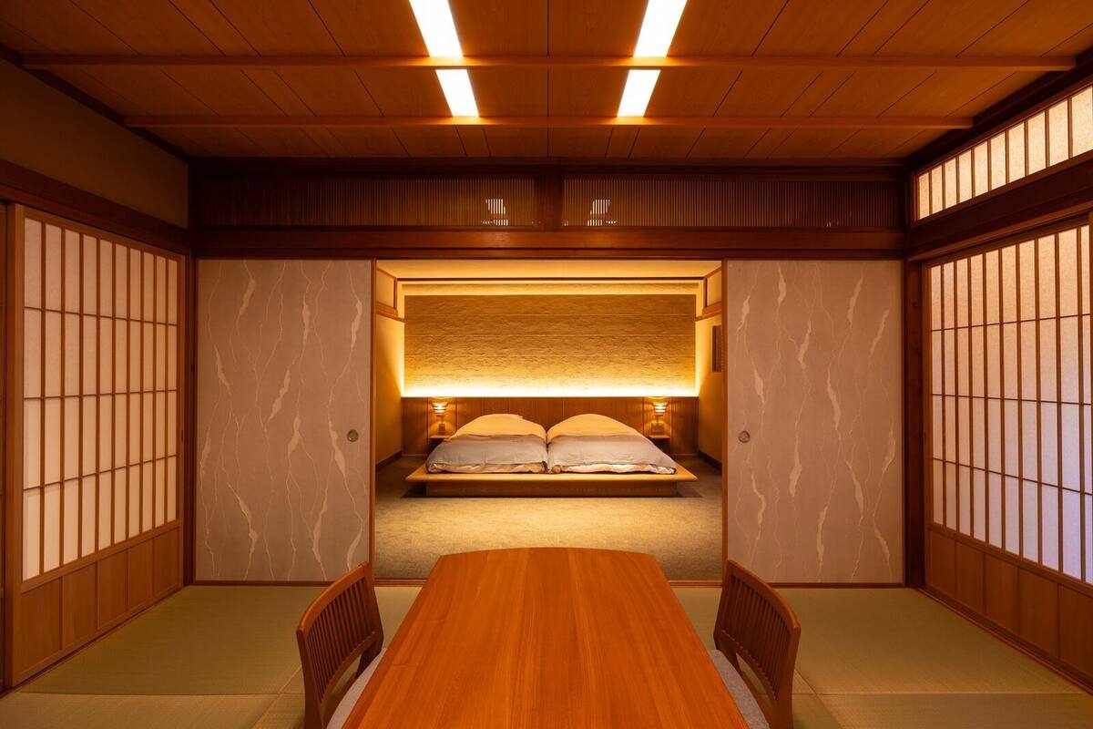 Une pièce de style japonais traditionnel, avec un sol recouvert de tatamis, un lit bas en bois doté d’une tête de lit et une table en bois avec des chaises.
