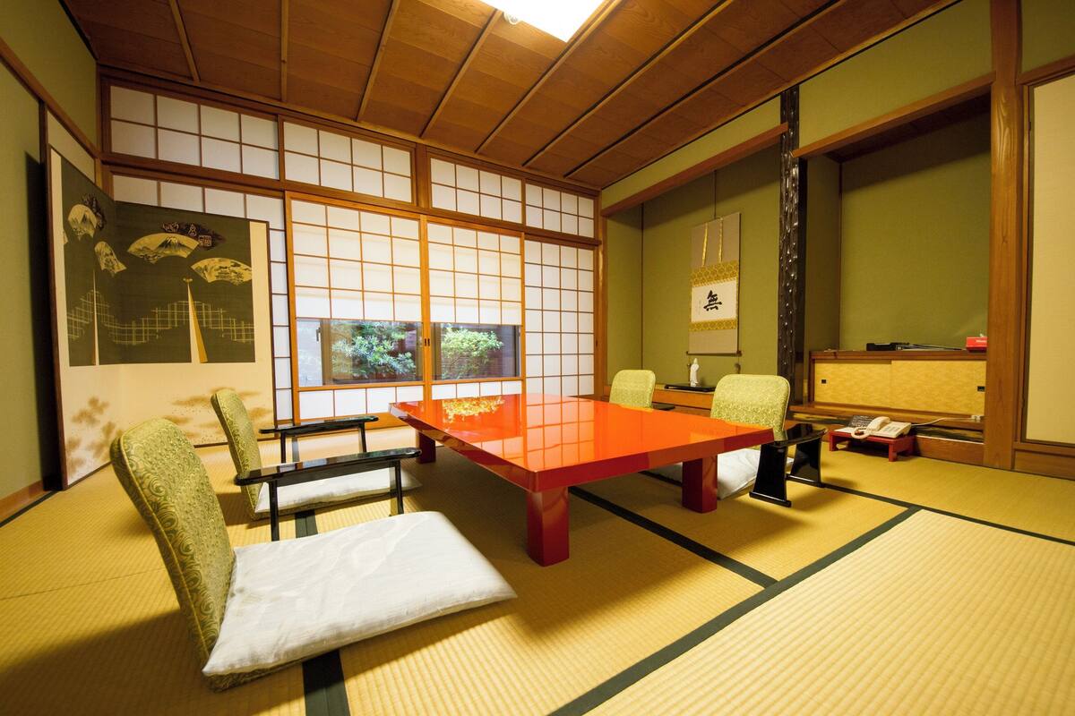Une pièce au sol recouvert de tatamis, avec une petite table basse, des chaises, un coussin, une fenêtre donnant sur un beau paysage et un paravent japonais traditionnel.