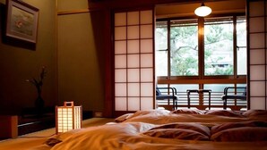 A traditional Japanese-style room with tatami flooring, a low bed with a futon, a shoji screen door, and a view of greenery outside. - Mikuniya (Toyooka)
