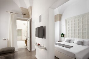Un dormitorio moderno con una cama grande, un televisor montado, un cabecero decorativo y una pequeña estantería con objetos.