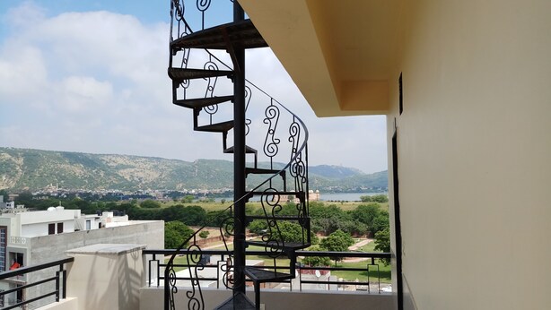 A spiral staircase with ornate metal railings leading to a balcony overlooking a scenic landscape with hills and a body of water. - Royal View Hotel (Jaipur)