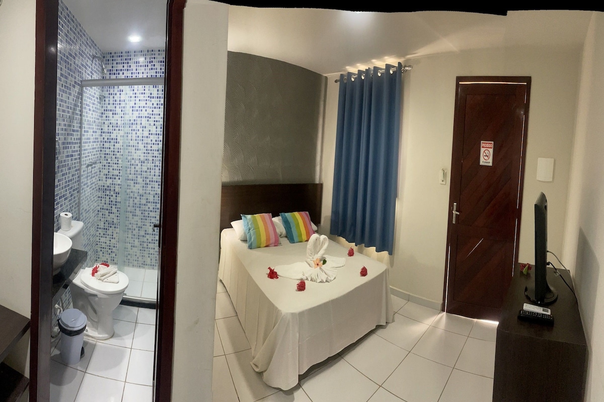 Baño con ducha, inodoro y lavamanos. Se ve una cama con cubrecama blanco y almohadas de colores. También se observa una mesita con mantel blanco y flores rojas.