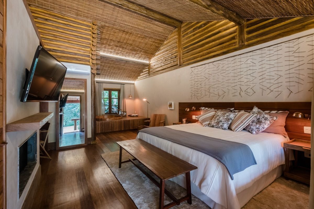 A bedroom with a large bed, a wooden bench, a TV, and a fireplace.