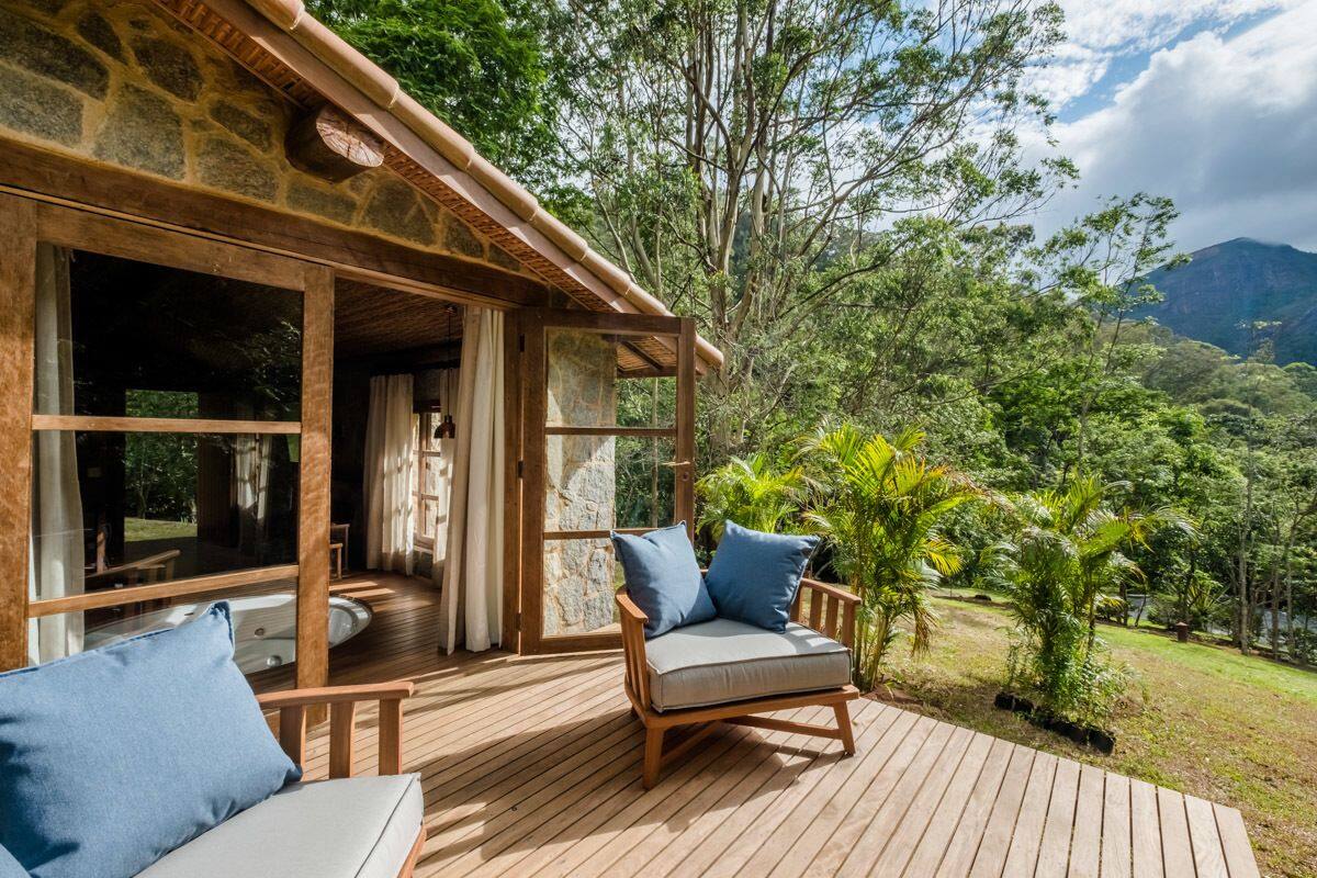 Una terraza de madera con un sofá y un sillón, un edificio de piedra y vistas a un paisaje exuberante con árboles y una montaña.