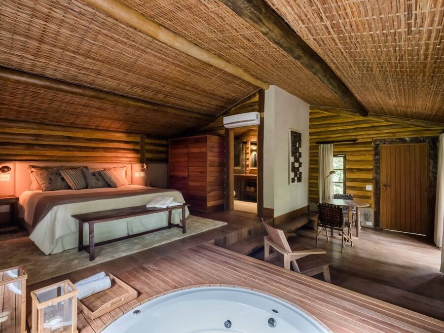 A cozy log cabin bedroom with a large bed, a wooden bench, a hot tub, and a dining area with a table and chairs.