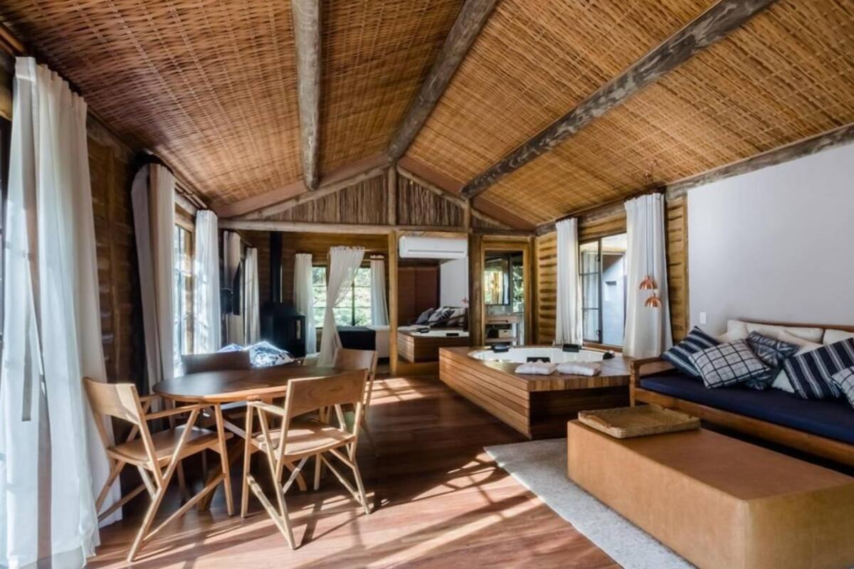 Una habitación amplia con techo de madera, un área de comedor con sillas y una mesa, un sofá con cojines, y un gran ventanal que proporciona luz natural.