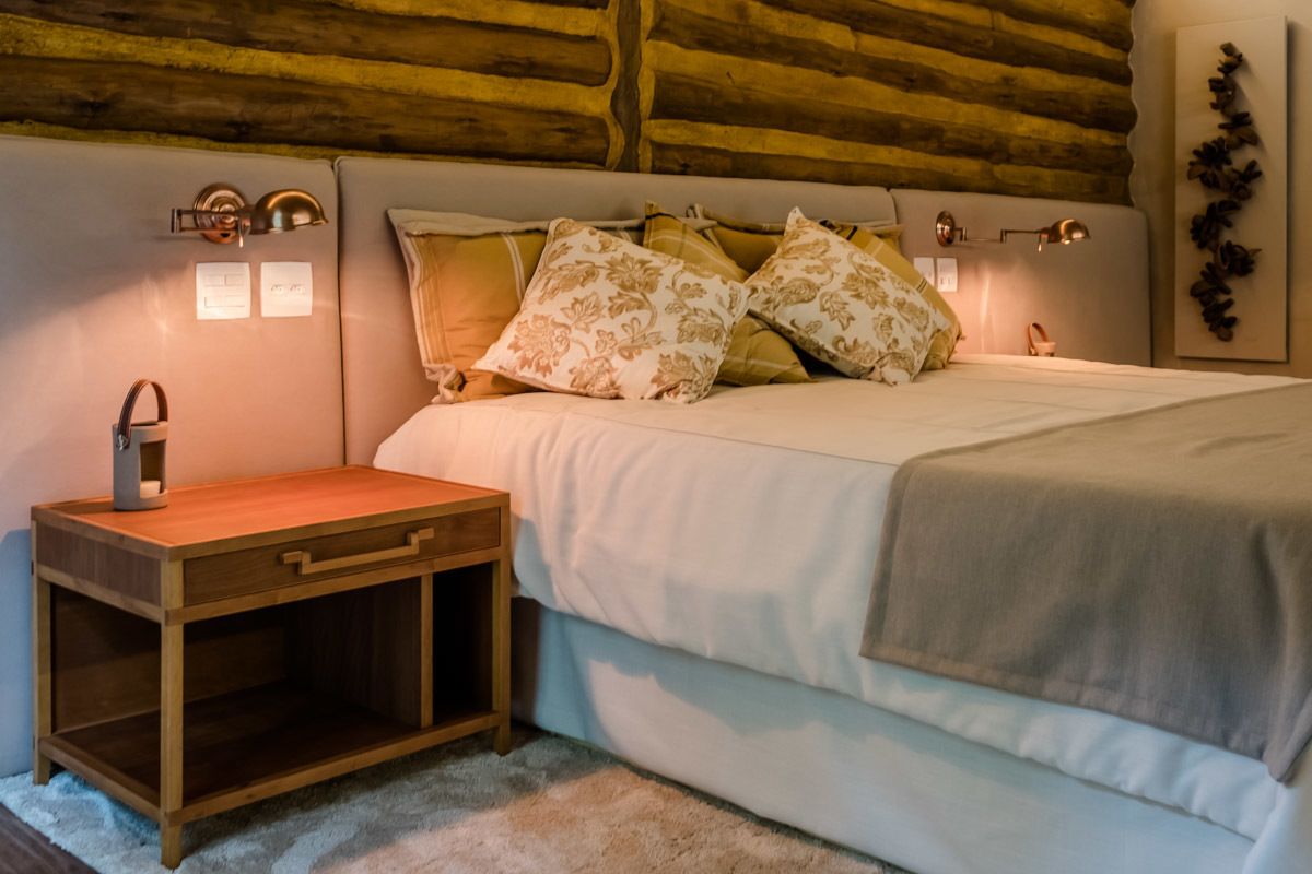 Quarto com cabeceira de madeira, mesa de cabeceira, abajur e cama com almofadas estampadas.