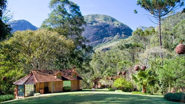 A rustic log cabin with a tiled roof, surrounded by lush greenery and a mountainous backdrop. - Pousada Tankamana (Petrópolis)