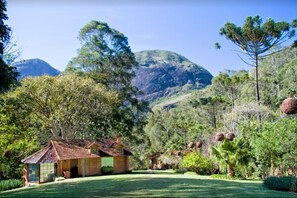 A rustic log cabin with a tiled roof, surrounded by lush greenery and a mountainous backdrop.