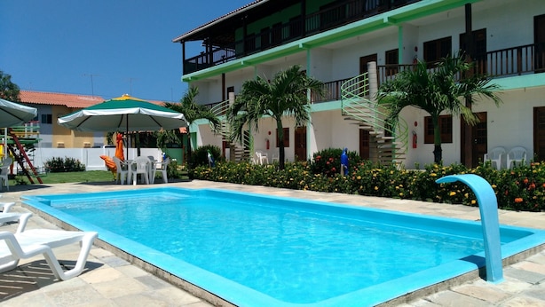 A swimming pool with lounge chairs and umbrellas, surrounded by a building with a balcony and stairs. - Pousada Porto de Galinhas (Ipojuca)