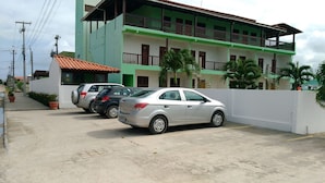 A multi-story building with a green and white facade, a white wall with a gate, and parked cars along the street. - Pousada Porto de Galinhas (Ipojuca)