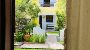 A view through a window of a courtyard with a pathway leading to a building with a blue and white facade. - Aguamarinha Pousada (Ipojuca)
