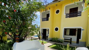 A courtyard with a yellow building, white benches, and a tree with pink flowers. - Aguamarinha Pousada (Ipojuca)