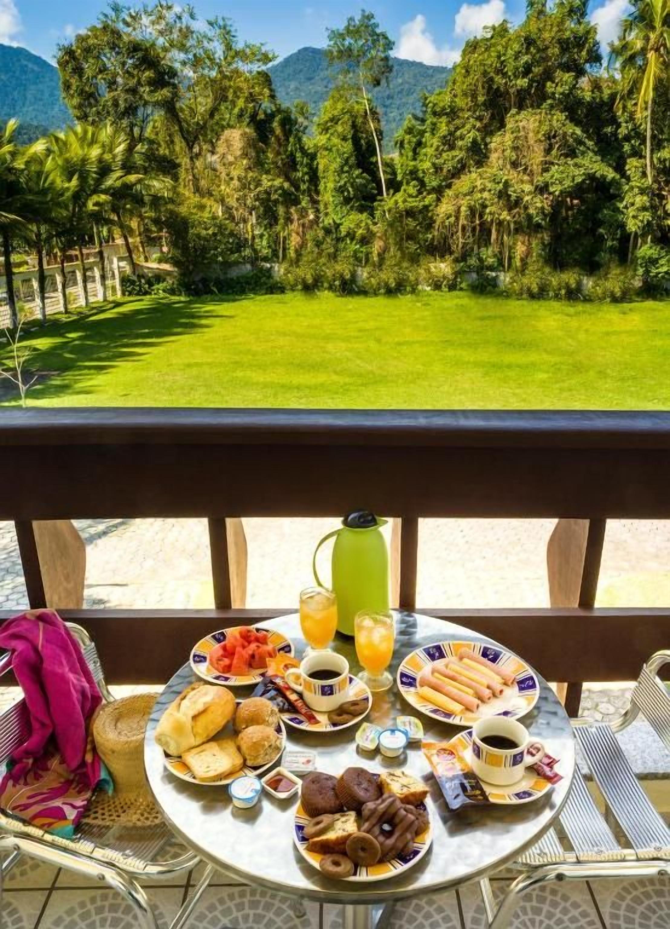 Una mesa de desayuno con diversos alimentos y una cafetera verde en un balcón con vistas a una zona cubierta de césped, árboles y montañas.