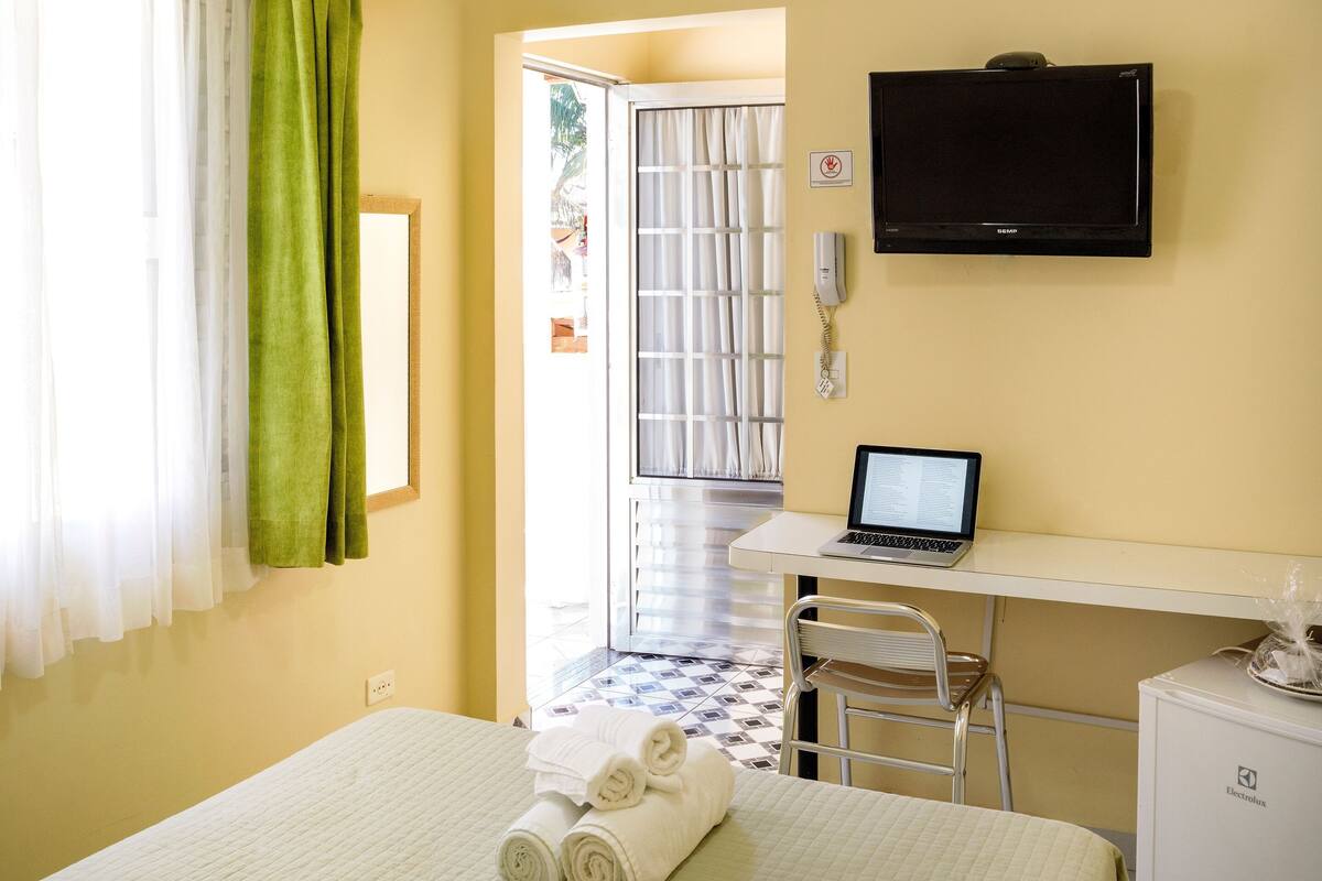 A hotel room with a bed, a desk with a laptop, a television, and a small refrigerator.