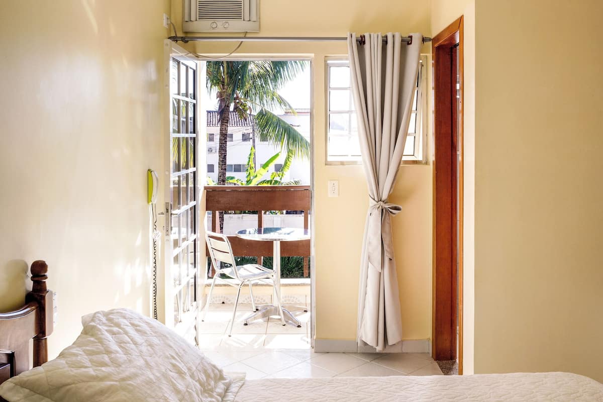 A bedroom with a bed, a window with curtains, an air conditioning unit, and a view of a balcony with a table and chairs.
