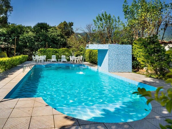 Una piscina rectangular con bordeado de azulejos, rodeada por una terraza también de azulejos. Hay sillas blancas y una pequeña mesa cerca de la piscina.