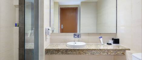 A modern bathroom with a granite countertop, a white sink, a glass shower enclosure, and a wooden door in the background.