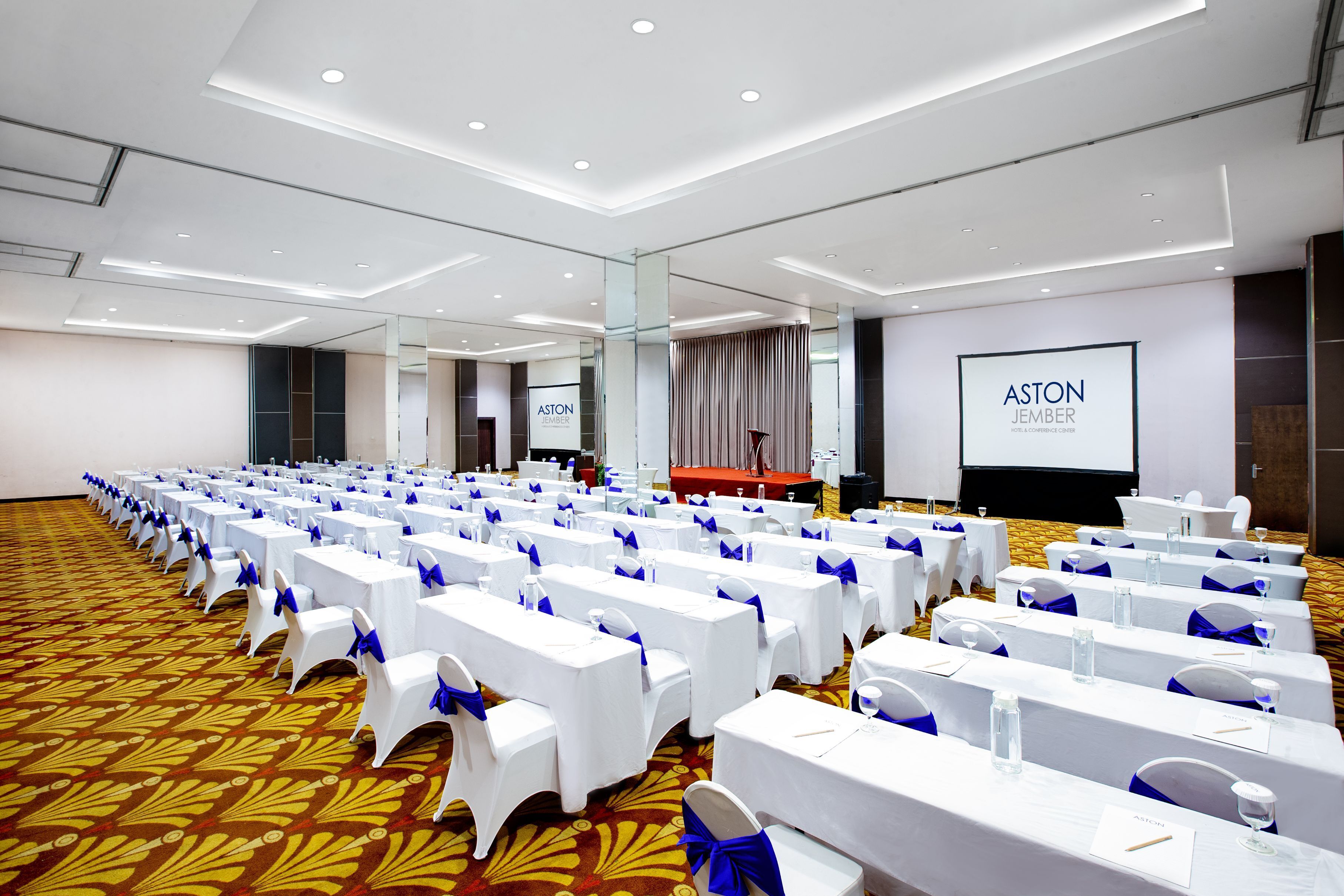 A conference room with rows of white tables and chairs, a podium, and a screen displaying 'ASTON JEMBER'.