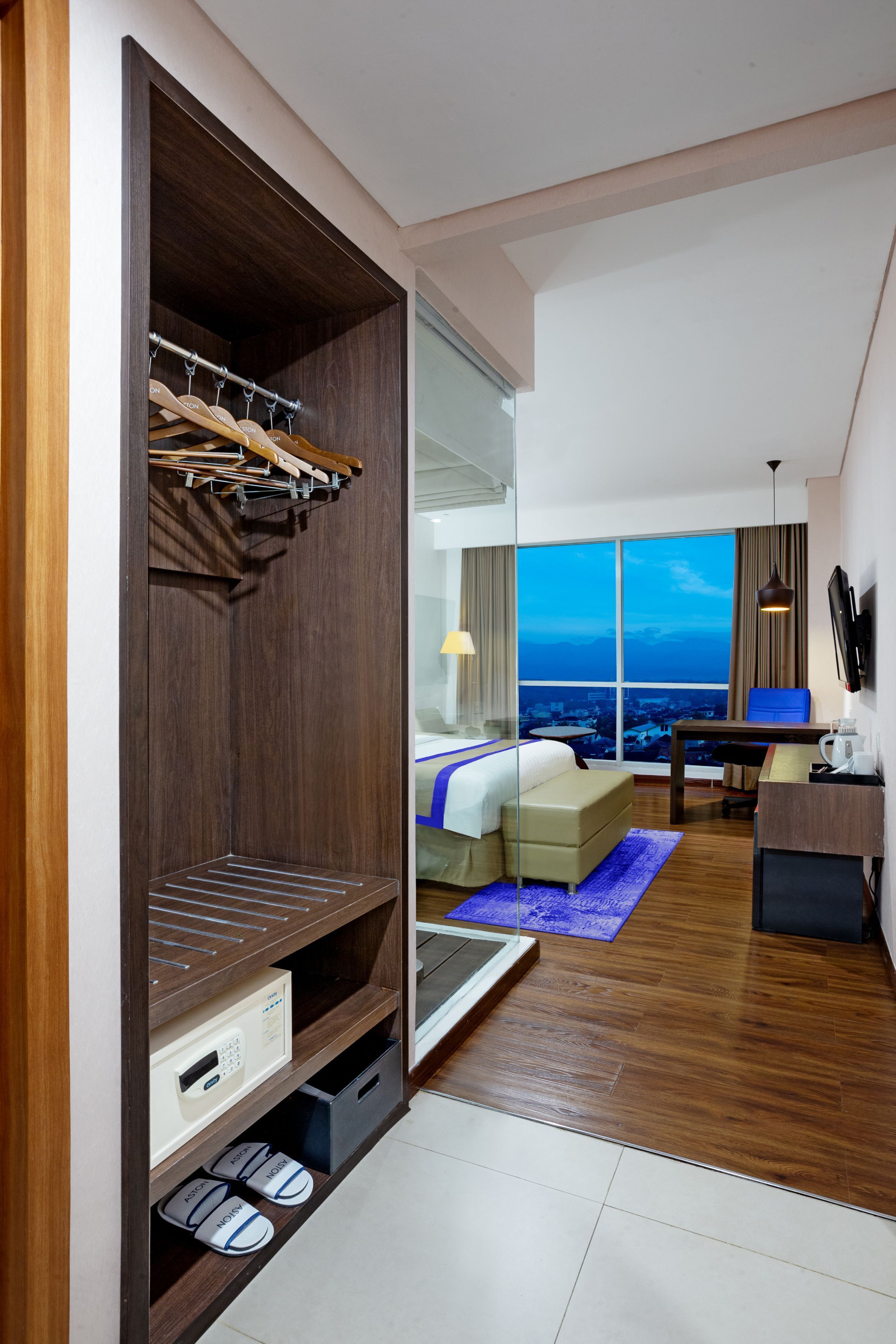 A hotel room with a wooden wardrobe, a bed with a blue accent, a desk with a chair, and a large window offering a city view.