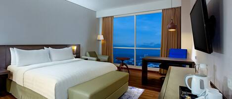 A hotel room with a large bed, a desk, a television, and a view of the city at dusk.