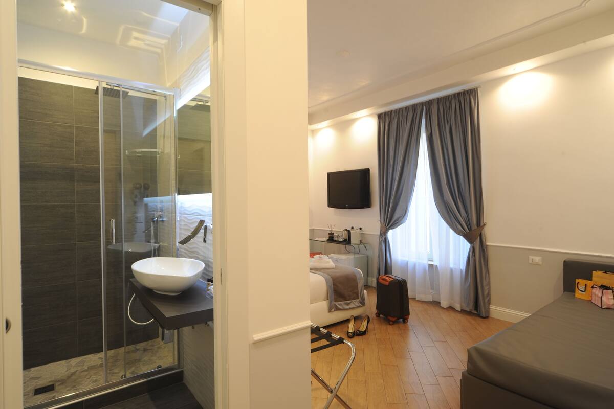 A modern hotel room with a glass-enclosed shower, a large bed, a bedside table with a flat-screen TV, and a window with curtains.