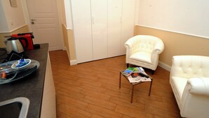 A living room with a white armchair, a small table with magazines, a coffee table, and a kitchen area with a kettle and a kettle on the counter. - My Suites Piazza di Spagna (Rome)