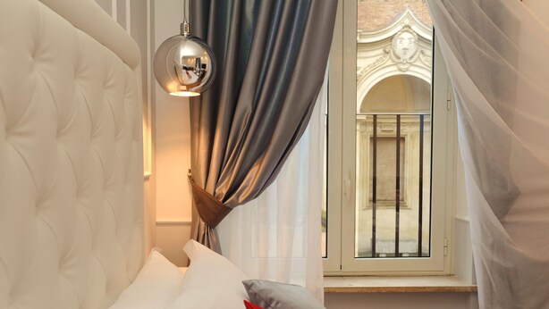 A bedroom with a tufted headboard, a bedside lamp, a window with curtains, and a view of a building through the window. - My Suites Piazza di Spagna (Rome)